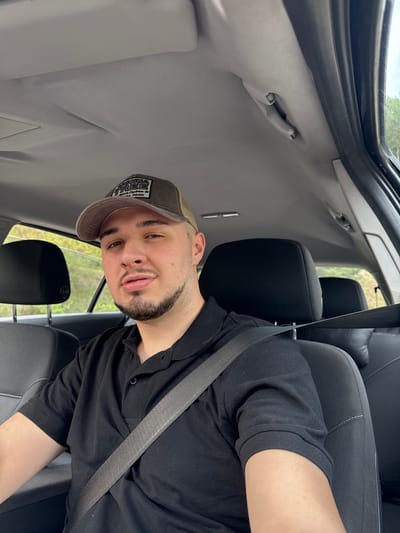 A young man is seated in the driver's seat of a vehicle, wearing a black polo shirt, a cap with an emblem, and a fastened seatbelt. The interior of the vehicle is visible, including the seats and ceiling. The background, visible through the car window, shows a glimpse of greenery. The man is looking towards the camera. There is no visible evidence to suggest that the location is the city of Byala Reka, Bulgaria.