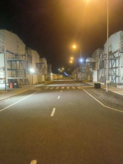 The image depicts a nighttime scene on a paved road, likely within an industrial or utility complex, as suggested by the presence of large, white, modular structures with external metal staircases and railings lining both sides of the road. Streetlights illuminate the area with a warm, yellowish glow, and some overhead lighting fixtures are visible. The road has white lane markings and a crosswalk. On the left side of the road, near one of the buildings, are three trash bins, two red and one green. A dark green, box-like object, possibly a generator or electrical enclosure, sits on a gravel area on the right side of the road. The environment appears to be undeveloped or sparsely populated, with a dark sky overhead. The overall impression is one of an operational facility at night, possibly in a remote or industrial zone. Based on the prompt, this scene is identified as being in the rural district of Qosshaghyldy, Qosshaghyldy aýyldyqOKRÝGİ, Kazakhstan. There are no people or animals visible in the image.
