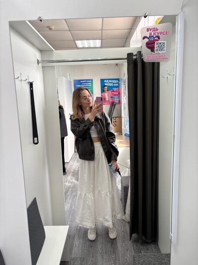 A woman stands inside a fitting room in a store, taking a selfie in the mirror with her phone. She wears a white top, a long white skirt, and a black jacket. In her other hand, she holds a water bottle. The fitting room has a gray wood-like floor, a white bench and walls, and a black curtain. A sign above the curtain has the text "БУДЬ В КУРСЕ!" and "ПОДПИСЫВАЙСЯ НА ТЕЛЕГРАM OZON", along with a QR code and a cartoon character. Other advertisements and signs are visible on the wall behind her.