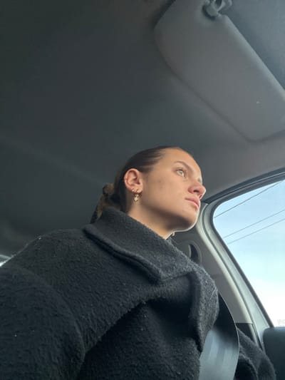 A young woman in a car looks upwards through the sunroof. She wears a dark wool coat and a seatbelt. Her hair is pulled back, revealing small, snowflake-shaped earrings. The car's interior is visible, including the sun visor and window frame. The sky is overcast and a telephone wire is visible through the sunroof. The image is taken from a low angle, emphasizing the woman's upward gaze.