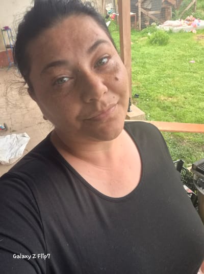 A person is captured in a close-up, slightly angled selfie, set outdoors in Kalipetrovo, Bulgaria. The subject, likely a woman, has dark hair pulled back, prominent green eyes, and a fair complexion with visible freckles or skin texture on her face. She wears a plain black short-sleeved top and looks directly at the camera with a neutral to slightly smiling expression.

The background suggests an outdoor, possibly rural or backyard setting. Immediately behind the person is a wooden railing or beam. Beyond this, a vibrant green grassy area is visible, indicating daylight and likely clear weather. Further in the background, elements like a wire fence, a small blue and red metal frame, and some white fabric or debris can be discerned. The ground directly behind the subject and to her left appears to be a paved or concrete surface.

A distinct watermark "Galaxy Z Flip7" is visible vertically on the black fabric of her top, indicating the device used to capture the image. The overall impression is a casual, personal photograph taken outdoors.