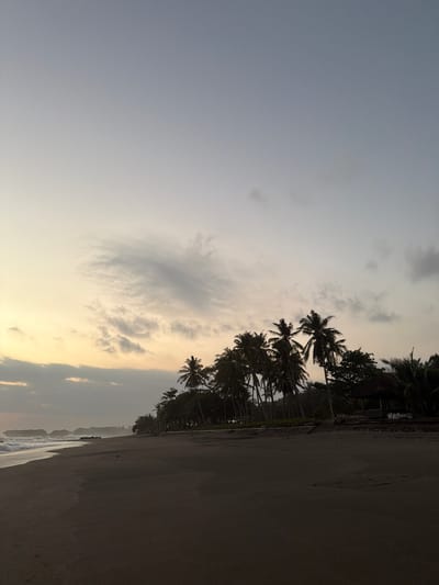 The image displays a wide-angle view of a dark sandy beach at dawn or dusk in Selemadeg Timur, Indonesia. The sky, occupying the upper two-thirds of the frame, exhibits a gradient from a pale yellow-orange at the horizon to a light blue-grey overhead, punctuated by scattered, wispy grey clouds. A singular, faint bright point, possibly a celestial body, is visible in the upper left portion of the sky. The ocean stretches along the left side, featuring small waves breaking onto the shore. In the far left distance, multiple dark rock formations or small islands are present in the water. A wide expanse of dark, wet sand covers the foreground and middle ground of the beach. Along the right side, a dense line of silhouetted palm trees and other tropical foliage forms a natural boundary. Within this tree line, a dark, thatched-roof structure, likely a gazebo or hut, is visible towards the right. Faint artificial lights are discernible behind the distant tree line. No individuals or prominent man-made objects are present on the immediate beach.