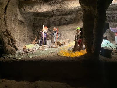 This media file depicts an underground scene, possibly a mine or cave, in Tolk, Germany. The image is oriented upside down, with the "ceiling" of the scene appearing at the bottom of the frame and the "floor" at the top. The setting is rough and textured, resembling a cave wall with prominent rocky formations and dirt.

The primary subjects are three gnome-like figures, appearing to be animatronic or puppet figures, engaged in some form of activity. Each gnome is dressed in distinctive clothing. One gnome wears a grey outfit and holds a red tool. Another is clad in overalls and a striped shirt, holding a wooden pole. The third, partially obscured, wears a green cap and a lighter-colored outfit. Their facial features suggest a friendly or industrious demeanor.

The scene is illuminated by artificial light sources. Clusters of translucent, colorful objects, resembling large crystals or glass shards, are embedded in the "ceiling" of the cave. Some of these are yellow and others are pale green, casting a warm and cool glow respectively onto the rocky surfaces below. The figures appear to be positioned as if working or performing a task within this environment.

Notable details include the rough texture of the cave walls, the variety of colored "crystals" providing ambient lighting, and the upside-down orientation of the image, which creates a unique visual perspective. The activity appears to be static, suggesting a display or diorama rather than a live event. There is no visible text in the image. The overall impression is one of a whimsical, subterranean display, possibly a tourist attraction.
