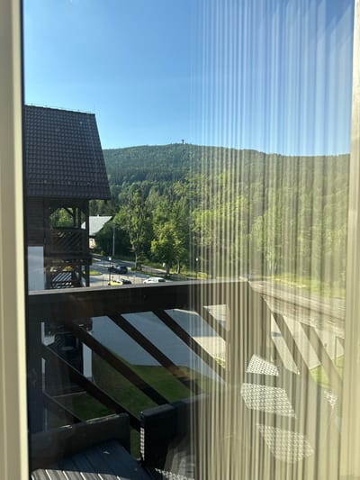 A view from a balcony in Szklarska Poręba, Poland, looks out onto a landscape of lush green hills under a clear blue sky. In the foreground, the wooden railing of the balcony is visible, with patterns from blinds casting vertical lines across the scene. To the left, a section of a dark-tiled roof and the wooden structure of a building are present. In the middle ground, a road runs through a clearing, with several cars parked and driving. Beyond the road, the hillside is covered in dense green trees, and a tall observation tower is visible on the crest of the furthest hill.
