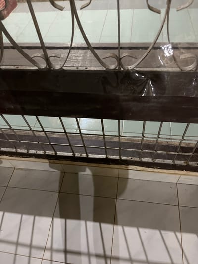The image depicts a close-up view of a window or door frame with decorative metal bars. The frame appears to be made of dark wood, and some sections are covered with dark plastic sheeting. Below the frame, there is a tiled floor, and shadows cast by the metal bars are visible on the tiles. The environment suggests an indoor or semi-outdoor setting, possibly a residential or commercial building. There are no people or discernible actions taking place in the image. The specific location in Kota Kuala Simpang, Indonesia, cannot be determined from the visual information provided.