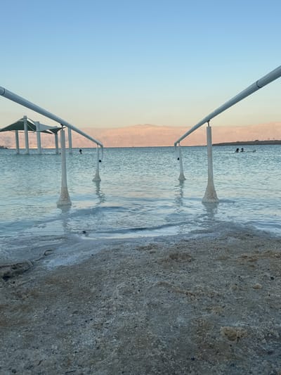 The image depicts a view of the Dead Sea at the Ein Bokek beach, located in the Tamar Regional Council, Israel. The foreground is a beach covered in coarse sand and salt deposits. In the middle ground, the Dead Sea water is visible, with structures of metal railings supported by pillars that are encrusted with salt deposits. The water is calm and slightly opaque, with some areas of white salt foam along the shoreline. In the background, there are distant mountains and the sky is clear and pale blue, indicating either early morning or late afternoon light. There are several people in the water, appearing to be swimming or floating due to the buoyancy of the Dead Sea. Two white shaded canopies are visible in the water further back. The scene captures a typical view of a tourist area at the Dead Sea.