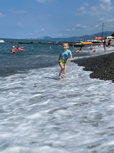 A young boy in a blue and green swimsuit runs through shallow waves at a pebble beach in Khostinskiy, Russia. The boy is mid-stride, with his arms extended and his mouth open in a happy expression. White foam from the waves surrounds his legs and feet. In the background, other people are swimming and relaxing in the sea. A long, dark breakwater extends into the water, with buildings and mountains visible on the distant coastline under a blue sky with scattered clouds. A yellow and red inflatable boat and a green sailboat are visible on the water near the shore.