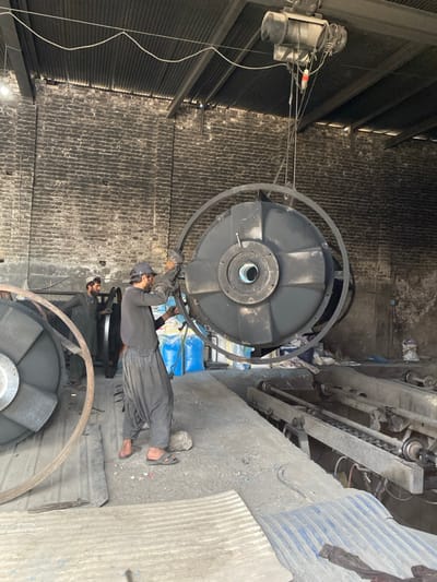This image captures an industrial workshop scene in Daman, Afghanistan. Two men are present within a large building featuring dark, rough-hewn brick walls and a corrugated metal roof. An overhead electric hoist system with multiple cables suspends a large, dark-colored circular industrial fan or impeller housing, which is encircled by a prominent metal ring. In the foreground, a man wearing dark clothing, a dark cap, and sandals is actively engaged with the suspended component, appearing to adjust or work on it. To the left, a second man in lighter attire and a white head covering stands near another similar, stationary large circular metal component. The floor is uneven, composed of dirt and concrete, with scattered debris, rocks, and corrugated metal sheets. Blue industrial sacks are visible in the mid-background, while a floor-level mechanical system with rollers and rails is partially visible on the right.