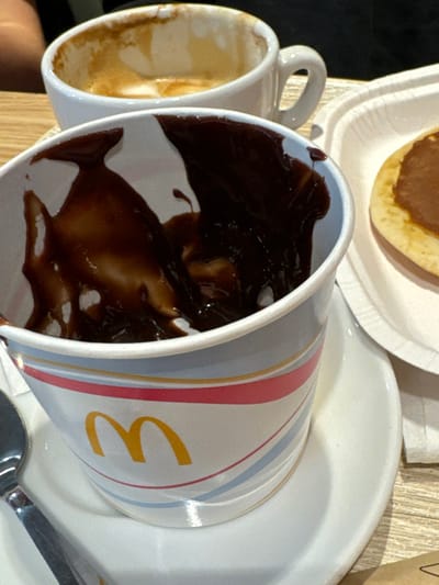 The image captures a close-up, slightly elevated perspective of a table within a McDonald's restaurant in Lipomo, Italy. Prominently in the foreground, a white paper cup displays the yellow McDonald's "M" logo and horizontal stripes in blue, pink, and yellow. The interior of this cup is coated with a thick, dark brown, viscous residue, consistent with melted chocolate or chocolate sauce. The cup rests on a white ceramic saucer. To the lower left, a portion of a silver spoon is visible. In the background, slightly left of center, a white ceramic coffee cup, approximately one-quarter full, contains light brown liquid with residual foam and staining, indicating a consumed coffee beverage. To the right, a segment of a light brown, circular food item, resembling a pancake topped with a spread, is visible on a white disposable paper plate. All items are situated on a light-colored, textured surface, indicative of a wooden or laminate tabletop. No individuals are visible in the frame.