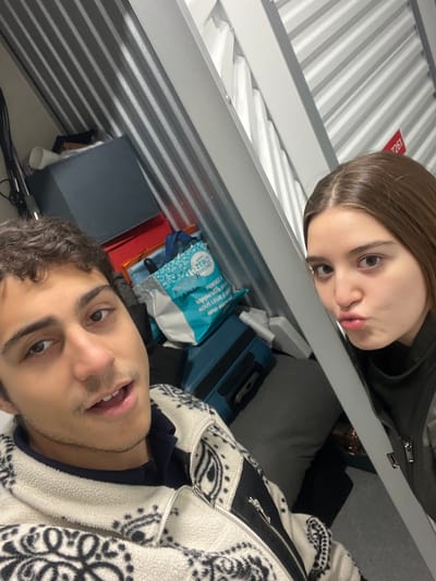 Two individuals are visible in the image. One individual, a female with light brown hair, is positioned above. Her lips are pursed. The second individual, a male with curly dark hair, is positioned below and is looking up into the camera.
The background includes luggage, including a blue bag and a blue suitcase, and some other items on the storage space above. The surroundings suggest the inside of a train. The presence of train carriage construction details suggest this may be a scene from a train journey.
Based on the context cues of location tagging and the presence of train construction and cabin details, this could be a train journey within Malakoff, France. However, there isn't any definite feature that explicitly ties this image location.