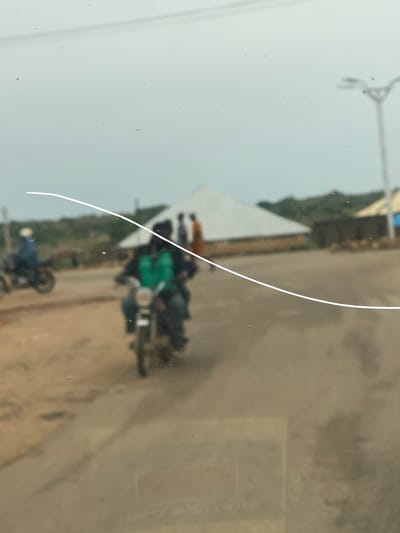 The image depicts a street scene in an urban area, likely DU, Nigeria, characterized by its dusty terrain and modest structures. In the foreground, two individuals are riding a motorcycle, with the rider in the front wearing a bright green vest. Behind them, another motorcycle with a single rider, wearing a blue shirt, is visible on the left side of the frame. In the background, several buildings with white, pointed roofs are present, along with other structures and vegetation. A lamppost stands on the right side of the image. The sky is overcast, suggesting a cloudy day. The image appears to be taken from a vehicle, as indicated by a horizontal white line, possibly a scratch on a window, that runs across the frame. The overall impression is one of a simple, everyday scene in a developing urban environment.