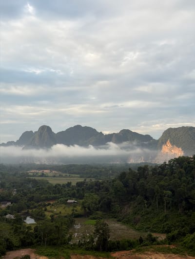 This media file captures a serene landscape scene in Hong Ngern Village, Laos. The image is oriented vertically, presenting a view of lush green mountains in the background, partially obscured by a gentle mist or fog. These mountains are characterized by their steep, rocky slopes, some of which are bare and show signs of erosion, while others are densely covered with tropical vegetation.

In the foreground and mid-ground, a dense forest blankets the land, interspersed with small human settlements. These settlements appear to be simple villages with scattered houses, some with thatched roofs. Patches of agricultural land, likely rice paddies, are visible, showing the coexistence of nature and human cultivation. A small body of water, possibly a pond or a slow-moving stream, adds to the tranquil atmosphere.

The sky is overcast with soft, diffused light, suggesting it could be early morning or late afternoon. The overall mood of the image is peaceful and somewhat mystical, enhanced by the presence of mist that softens the distant features. There are no visible people or specific activities depicted; the focus is on the natural beauty and the quiet life of the village. The scene evokes a sense of remoteness and a connection to nature. No text is visible in the image.