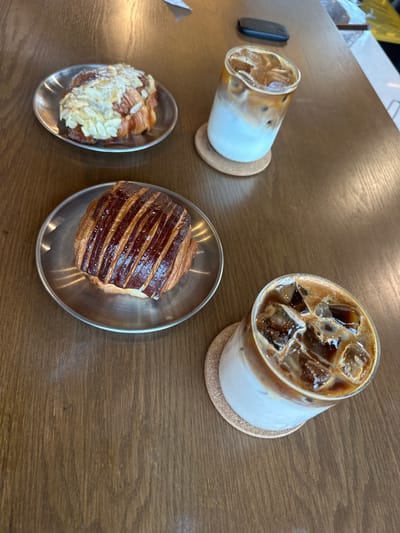A high-angle, medium-shot photograph shows a wooden table with two glasses of iced coffee, two pastries, and a black object. The coffee drinks are in cylindrical glasses with ice, coffee, and a white liquid (likely milk). Both are resting on small, round, cork coasters. The pastry closest to the lower left is an almond croissant, while the other is a plain croissant with glazed, striped dark brown topping. Both pastries are displayed on small, oval, metal dishes. To the left of the lower coffee drink is a black, rectangular object. The scene is possibly set in a cafe or restaurant in San Pedro Garza García, Mexico. No people are visible in the image.