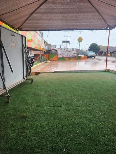 The image depicts a street scene in Bwari, Nigeria, under an overcast sky, suggesting it might be raining or recently rained as the ground appears wet and reflective. In the foreground, a patch of artificial green turf is visible, bordered by a concrete edge. To the left, a large, metallic structure, possibly a part of a stall or storage unit, is positioned on wheels, indicating it might be movable. The structure has a weathered appearance with some discoloration and visible hinges.
Beyond the turf and the metallic structure, a building with a vibrant facade featuring a checkered pattern of orange and green panels is visible. A banner with text and images, seemingly related to a training program, is hung across a fence or railing in front of the building. A light-colored sedan is parked on the wet road in front of the building, partially obscured by the banner. Overhead, a canopy structure made of a dark, textured fabric is supported by poles, casting a shadow over the scene. In the background, more buildings and trees line the street, hinting at a typical urban or semi-urban environment. The overall impression is a slightly damp, commercial or institutional area.