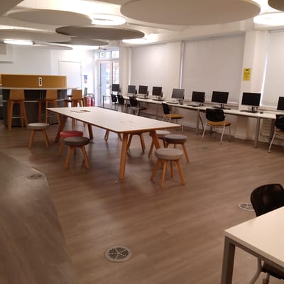 This media file captures a modern, empty study or computer lab area within an institutional building, likely in Coventry, United Kingdom. The room is brightly lit, featuring several large, circular ceiling lights and acoustic panels suspended from above.
The space offers various work and seating arrangements. Along the right wall, a series of white desks are equipped with black computer monitors, keyboards, and mice, accompanied by black office chairs with either yellow or grey seats. The windows along this wall are covered by white roller blinds. A yellow sign displaying "CCTV" and a camera icon is visible on a pillar, indicating surveillance.
In the center, a long, light-colored wooden table with angled wooden legs is surrounded by several round stools with upholstered tops in shades of grey, red, and beige. Towards the left, a tall, semi-circular counter with wooden bar stools provides a more casual workspace, featuring power outlets on its surface.
The flooring is a consistent light brown, wood-effect material, punctuated by several circular metallic grates. A glass door on the far left provides a view of an outdoor area with trees and buildings, and a red fire extinguisher is positioned nearby. Another white door is also visible. The absence of people suggests the image was taken during off-peak hours. The overall environment appears clean, organized, and functional, designed for study or collaborative work.
