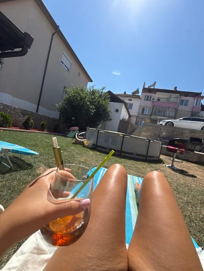 A first-person perspective shot captures a person lounging on a sun-drenched lawn in Sandanski, Bulgaria, on a bright, clear day. The individual is reclined on a striped beach towel, with their tanned legs prominently displayed in the foreground. Their right hand holds a glass filled with a dark amber liquid and ice, with a green straw and a white cylindrical object, possibly a vape pen, resting inside the glass. The background reveals a residential area with several buildings of varying heights and architectural styles, some featuring tiled roofs and solar water heaters. A large, above-ground swimming pool is visible in the middle ground, surrounded by a lush green lawn and scattered small bushes. In the distance, a white sedan is parked on an elevated concrete platform. The overall scene depicts a leisurely summer day outdoors.