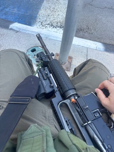 A close-up, overhead view depicts the lower body of an individual, clad in olive green tactical pants, seated outdoors in the Brener Regional Council, Israel. A rifle, consistent with an M4/AR-15 platform, rests horizontally across the lap. The rifle is equipped with a black heat shield on the handguard, a birdcage flash hider, and a black tactical accessory (likely a light or laser) mounted on the front. A green paracord is tied near the front sight post. The rifle's charging handle displays an orange safety tag. A black tactical sling is visible across the person's left thigh, and a portion of a green tactical pouch is seen on the lower left. The individual's right hand rests on the rifle's upper receiver, near the bolt carrier group. The ground consists of concrete or asphalt, alongside a grey metal pole anchored by four bolts, and a curb painted blue and white. A circular metal utility cover is partially visible on the ground.