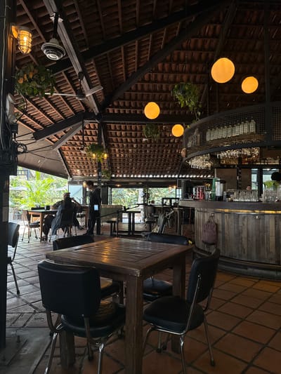 The image depicts the interior of a restaurant, possibly in La Tijera, Mexico. The scene is taken from an unusual, near-vertical perspective. Dominating the view is the restaurant's high, angular roof, constructed with dark metal beams and terracotta tiles, which create a visually textured pattern. Hanging from the roof structure are several round, warm-toned light fixtures.
Below the roof, the restaurant features a wooden bar area stocked with glassware and bottles. A tall wooden table, surrounded by black chairs, occupies the center of the frame. Several people are visible, engaged in various activities such as standing and seated at the table, with blurred figures suggesting movement. Outside the restaurant, greenery is visible through large windows. The floor of the restaurant is covered in terracotta tiles arranged in a grid pattern. Cables and what appear to be audio or video equipment are situated in the lower foreground.