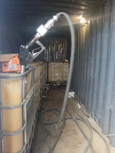 The image shows the interior of a shipping container in what appears to be an industrial setting. A row of large, caged, yellow/tan liquid totes lines the left side of the container, with a black fuel dispensing nozzle and hose positioned on top of the front totes. Cables, hoses, and electrical equipment are scattered across the floor. Other liquid totes are visible in the back of the container. The walls of the container are corrugated metal, lit by a single overhead light. The location is Яльгелево, Russia.