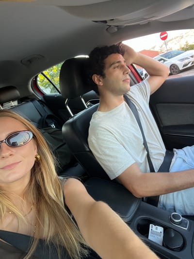 The image captures two individuals inside a red compact car, situated in Cacém e São Marcos, Portugal. The scene is illuminated by natural daylight.
In the foreground on the left, a female is depicted, likely taking a selfie. She has long, straight blonde hair, wears dark oval-shaped sunglasses, a dark-colored top, and gold hoop earrings. A delicate gold necklace is visible around her neck. Her left arm rests on the central console.
To her right, seated in the front passenger seat, is a male with dark, curly hair. He is wearing a light-colored short-sleeved t-shirt and light blue jeans. He is looking towards his right, out the passenger window, with his right arm bent and resting behind his head. Both individuals are secured by dark-colored seatbelts.
The car's interior features dark upholstery on the seats and door panels. On the center console, a black object resembling a small container or device, and a white paper with a barcode, are visible within a storage compartment. A dark backpack or bag is present on the rear seat.
Through the right-side passenger window, an international 'No Entry' road sign (red circle with a white horizontal bar) is prominent. Beyond the sign, parked vehicles, including a white SEAT, and buildings with light-colored facades and red tiled roofs, typical of the region, are observed. Green foliage is visible through the left rear window.