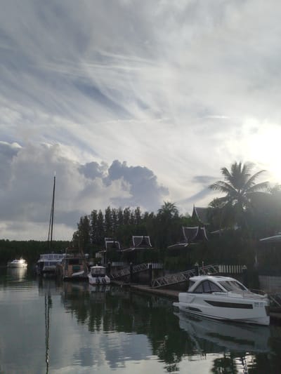 The image shows a canal or bay in Ko Kaeo, Thailand, with several boats docked. In the foreground, a modern white speedboat with the registration "DB 37" is prominently featured, reflecting in the water. Behind it, other boats, including a larger yacht and a sailboat, are visible. The shoreline is lined with lush trees and traditional Thai-style buildings with dark, pointed roofs. A tall palm tree stands out against the sky on the right side of the image. The sky is dramatic, with a mix of bright sunlight and dark, streaky clouds. The water is calm, with reflections of the boats and the surrounding scenery.