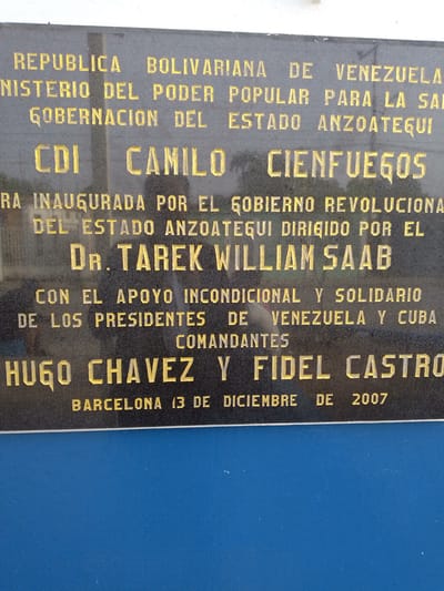 The image shows a plaque commemorating the inauguration of the "CDI Camilo Cienfuegos" in Barcelona, Venezuela, on December 13, 2007. The plaque is in Spanish and identifies several key figures associated with the project: the Bolivarian Republic of Venezuela, the Ministry of Popular Power for Health, the Governor of the State of Anzoátegui, Dr. Tarek William Saab, and Commanders Hugo Chávez and Fidel Castro. The plaque mentions the unconditional support and solidarity between the presidents of Venezuela and Cuba. The plaque is dark with gold lettering and is affixed to a blue surface.