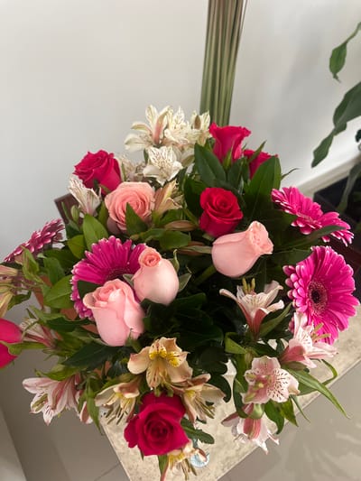 The image depicts a bouquet of assorted flowers, including pink gerbera daisies, light pink roses, and red roses, along with smaller white and pink alstroemeria flowers, arranged with green foliage. The flowers are placed on a surface, possibly a table. A wall is visible in the background. There are no people visible in the image. The location given is Barrio de Nuevo León, Mexico, though no specific information in the image confirms this location.