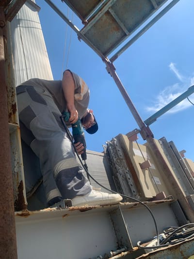 A man in work clothes is using a rotary hammer drill outdoors, under a clear blue sky. He is wearing a grey t-shirt with orange and white stripes on the sleeve, and grey work pants with darker grey panels and white trim. He has a dark blue cap on his head and is wearing glasses. The man is positioned on a metal structure, possibly a rooftop or scaffolding, with rusted metal beams and supports visible. In the background, there are large, light-colored panels stacked vertically. The scene appears to be a construction or industrial work area. There is no identifiable information that places this scene specifically in the city of Pivdennoukrainsk, Ukraine.