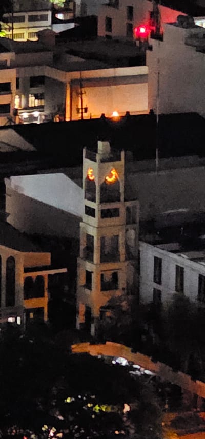 The image depicts a nighttime urban scene in Makati, Philippines. The focus is on the illuminated windows of several buildings, suggesting residential or commercial spaces. A traffic light with a red signal is visible on the left side, indicating a road intersection. In the center, a partially constructed or decaying building exhibits several open window frames, with warm orange light emanating from within some of them. These lights appear as abstract shapes, perhaps from interior fixtures or decorative elements. The overall impression is one of a bustling city at night, with a mix of active and dormant structures. The lighting suggests it is after sunset, and the absence of visible weather elements implies fair conditions. No text is discernable in the image.
