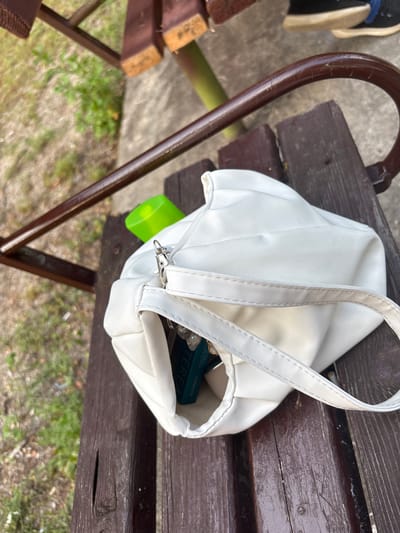 An overhead, medium shot captures a white handbag with a strap resting on a weathered wooden park bench in Garmen, Bulgaria. The handbag, made of a faux leather material, is partially open, revealing a bright green cylindrical object inside and what appears to be a book with a turquoise cover. The bench, showing signs of age with scuffs and a visible knot in the wood, is situated outdoors, with out-of-focus green foliage and a concrete path visible in the background. A dark brown, curved metal railing is also present, suggesting the bench is part of a public space or playground.