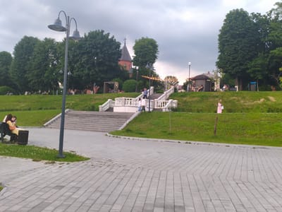 The image depicts a public park scene in Zavodskoy, Russia. A paved walkway with a herringbone pattern leads into the foreground. Beyond the walkway, a grassy hill slopes upwards. On the left side of the hill, two women are seated on a bench, one of whom is holding a purse. A lamppost with two globes stands prominently in the middle ground. Further up the hill, a grand stone staircase with decorative white balustrades ascends towards a gathering of people. Some individuals are interacting near the top of the stairs, and a woman in a pink dress walks along the path at the top of the hill on the right. In the background, a red brick building with a dark steeple, resembling a church or historic structure, is visible amongst lush green trees. A small, hexagonal structure with tables and chairs is situated to the right of the stairs. The sky is overcast.