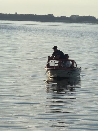 The image shows two people in a small boat on a body of water, possibly a lake or river. The boat has a red cabin with a glass windshield. One person, wearing a cap, is leaning over the side of the boat, appearing to be tending to something in the water or near the hull. The other person is sitting inside the cabin, looking out. The water's surface is disturbed, creating a rippled effect that reflects the light, suggesting it is a calm day with a slight breeze. The background shows a dark, tree-lined shore on the left side of the image, indicating a natural setting. The lighting suggests it is either early morning or late afternoon, with the sun casting long reflections on the water. There is no visible text in the image. The overall scene evokes a sense of quiet activity, perhaps fishing or leisurely boating.