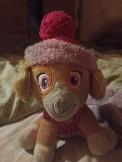 This is a close-up, eye-level, static shot of a plush toy dog, likely Skye from Paw Patrol, resting on a textured surface that appears to be bedding or fabric. The toy is facing forward, with its head slightly tilted. It is wearing a pink fuzzy hat with a red pom-pom on top and a pink vest. The toy's fur is light brown and tan, with white accents on its paws and muzzle. Its large, pink-rimmed eyes with black pupils are prominent. The background is out of focus, showing dark, indistinct shapes that could be furniture or a wall. The lighting is dim, creating shadows and highlighting the textures of the toy and its surroundings. There is no indication of the city of Berezivka, Ukraine, or any specific geographic location within the image.