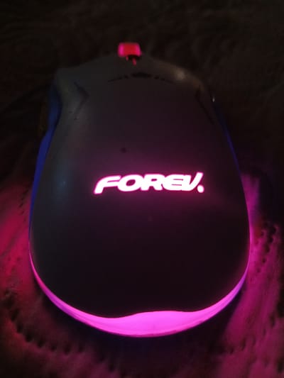 A gaming mouse with pink and blue lighting is positioned on a dark, textured surface, likely a mousepad or fabric. The mouse is dark gray and features a luminous pink logo that reads "FOREV." The logo is centered on the top of the mouse. The base of the mouse also emits a soft pink glow. The image is taken from a slightly elevated, top-down perspective, with the mouse as the primary subject. The lighting suggests a low-light environment, with the mouse's illumination being the dominant light source. There is no indication of a specific location within Ashkelon, Israel.