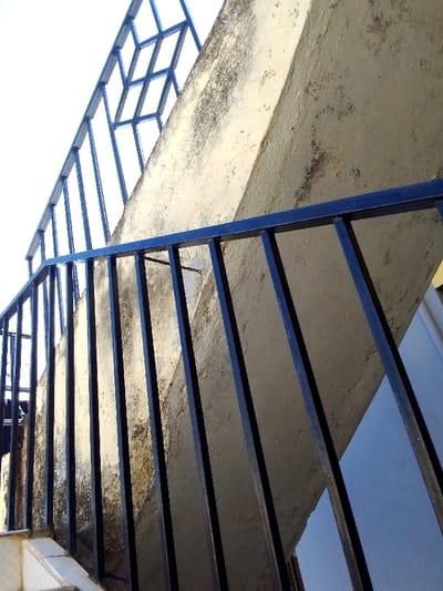 The image presents an upward-angle view of an exterior concrete staircase located in Kalimoni ward, Kenya. The stairs are constructed from light tan-colored concrete, exhibiting signs of weathering, surface discoloration, and localized dark staining consistent with dirt or biological growth. A dark-colored metal railing system runs along the left side of the staircase. The lower section of this railing, closer to the viewer, features a horizontal top rail supported by numerous vertical rectangular balusters. Further up and to the left, a segment of an upper railing is visible against a bright white sky, showcasing a more complex design incorporating diamond-shaped patterns within its metalwork. A small section of light-colored tiled flooring is visible at the bottom left, indicating a landing area. To the right of the concrete structure, a segment of a pale blue or light grey wall is visible. No individuals are present in the scene.