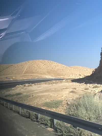 The image depicts a view from inside a vehicle, looking outwards. The scene is dominated by a clear blue sky with faint contrails from an aircraft, suggesting daytime. The vehicle's interior, with its reflective surface, takes up the left side of the frame. The right side of the image shows a rugged, arid landscape with a dry, sandy slope leading down to a road. A metal guardrail separates the road from the slope. The terrain is characterized by sparse vegetation, rocks, and patches of dry earth, indicating a desert or semi-arid environment. The lighting suggests it is daytime, possibly late afternoon given the angle of the sun. The image is oriented vertically, as if the camera was held sideways. There is no visible text in the image.