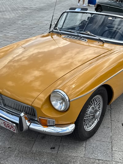 The image shows a close-up of a vintage yellow convertible car parked on a cobblestone street in Bruges, Belgium. The car, possibly an MGB, features a chrome grille, a round headlight, and a spoked wheel. The car's hood reflects the sky with scattered clouds. In the background, another dark-colored vintage car is visible, and two people are walking in the distance. The overall scene suggests a car show or a special event in Bruges.