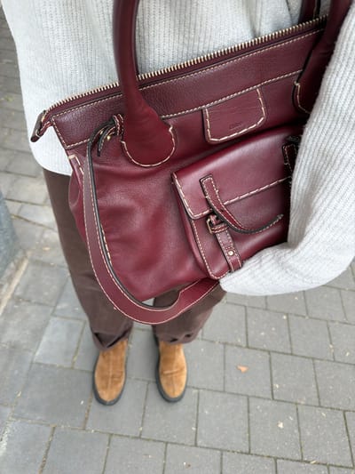 A high-angle shot shows a person standing on a gray brick walkway, holding a burgundy leather handbag. The person is wearing brown pants and tan suede shoes. The handbag has a zippered top and an outer pocket with a buckled strap. A white sweater is visible under the handbag. The background consists of the gray brick walkway. This could be Bad Sobernheim, Germany, based on the context.