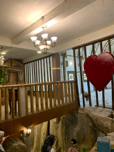 The scene is indoors in a building in Kecamatan Borobudur, Indonesia, located at latitude: -7.606, longitude: 110.18. There is a wooden walkway with bamboo railings. The walkway is suspended over a deep, rocky pit with a blue area in the lower right corner. There is a large red heart-shaped object hanging from the ceiling in the background, surrounded by a latticework structure. A chandelier hangs from the ceiling above the walkway. There is a wooden cabinet in the background and a small table with a wooden top under the walkway. There are several people in the scene, wearing head coverings, but their faces are not visible. The scene is lit by a single overhead light and the chandelier. The environment is relatively dark and appears to be a cave or an underground space.