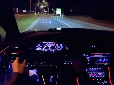 The image shows the interior of a car driving at night on a road that appears to be in or near Bezden, Bulgaria. The driver's hands are visible on the steering wheel, and the car's dashboard displays are illuminated, showing speed, gear selection, and other information. The road ahead is lit by the car's headlights and streetlights, with trees and buildings visible on the sides. The scene depicts a typical night-time drive, with the focus on the car's interior technology and the road ahead.