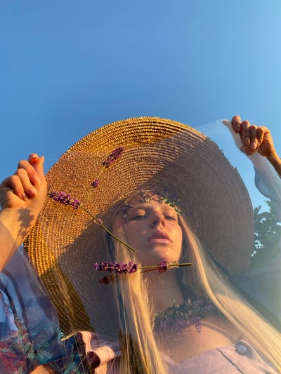The media file is a low-angle, close-up shot of a person's face viewed from below, reflected on a transparent surface against a clear blue sky. The person, a young woman with long blonde hair, wears a wide-brimmed straw hat adorned with small purple flowers, likely lavender, which also appear to be incorporated into a delicate floral crown around her head. Her eyes are partially closed, and her lips are slightly parted, conveying a serene or contemplative mood.

Her face is illuminated by soft, warm light, suggesting it is either early morning or late afternoon. The setting appears to be outdoors, with the clear blue sky as the dominant background element. A hand, with neatly manicured nails, is visible in the foreground, holding the transparent surface, which creates the reflection. Another hand is visible higher up, also interacting with the reflective surface. The person is wearing a light-colored top, possibly lilac, and parts of a shimmering blue fabric are also visible, hinting at clothing or a decorative element.

The overall impression is one of peacefulness and natural beauty, with the reflection adding an artistic and slightly surreal dimension to the image. The presence of lavender suggests a summery or pastoral atmosphere, possibly in a garden or meadow. No text is visible in the media file.