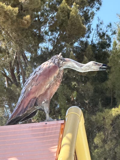 A bronze sculpture of a swan-like bird is mounted on the corner of a building. The building has salmon-colored vertical paneling and a yellow pipe running along the corner. The sculpture is positioned with the bird's head tilted upwards. The background consists of trees and a patch of blue sky. Based on the location information, this photo was likely taken in the city of Tías, Spain.