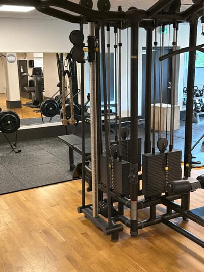 This image shows a gym in Östersund, Sweden. The main focus is a cable crossover machine with two weight stacks. In the background, a mirrored wall reflects rowing machines and other gym equipment. The floor is wooden, and a black rubber mat is visible in front of the cable machine. There are no people present in the image.