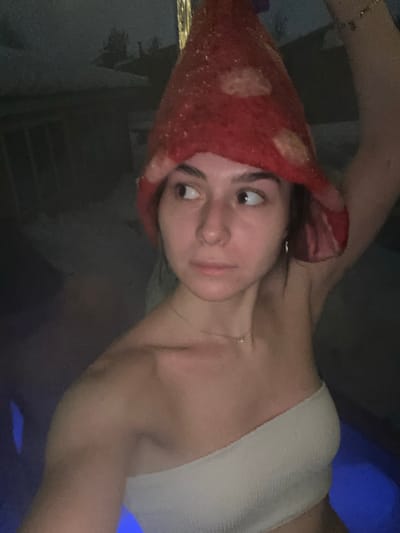 A female subject is depicted outdoors at night in Базарово, Russia, partially submerged in blue-lit water, likely a hot tub or pool. She wears a red, mushroom-shaped felt hat with light-colored spots and a light-colored, ribbed strapless top. A delicate gold-colored necklace and a silver-colored hoop earring are visible. Her dark hair appears damp, and her gaze is directed towards the left of the frame. Her right arm is raised, extending out of the frame at the top right. The background is dark and out of focus, showing indistinct silhouettes of structures and possibly trees under a dark sky.