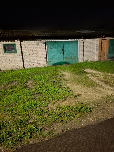 The image depicts a nighttime exterior view of several garage doors made of metal, painted a faded green, set within brick and cinder block walls. The garages appear to be part of an outdoor storage or parking facility, possibly in a residential or industrial area of Novlyanka, Russia. The ground in front of the garages is a mix of green grass and a gravel path, suggesting an unpaved or poorly maintained area. A small, barred window with a green frame is visible on the left side of one of the garages. The overall scene is dimly lit by artificial light, creating shadows and highlighting the weathered textures of the structures and the surrounding environment.