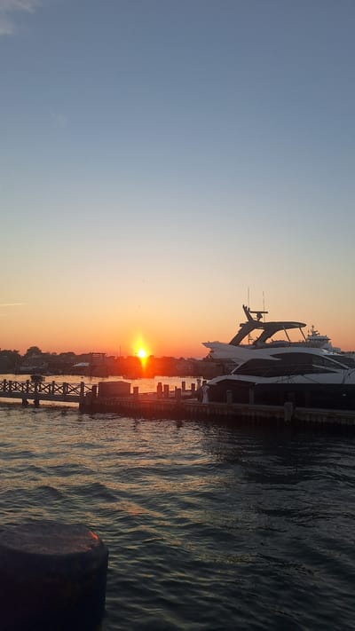 The image depicts a sunset over the waterfront in Nantucket, Massachusetts, United States. The sun is low on the horizon, casting a warm orange glow across the sky and reflecting on the water. Several yachts are docked at the pier, silhouetted against the vibrant sky. The foreground shows rippling water and a dark, cylindrical object, likely a bollard or mooring post, on the left. In the background, there's a wooden boardwalk with railings and a cluster of buildings. The overall scene is serene, capturing the tranquil beauty of a coastal sunset.
