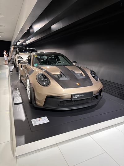 The image captures the interior of the Porsche Museum in Stuttgart, Germany. In the foreground, a golden-brown Porsche 911 GT3 RS is prominently displayed on a dark platform. The car features a carbon fiber hood with prominent vents and a black racing stripe. The front of the car has a black license plate holder with "PORSCHE MUSEUM" text. To the left of this car, a white Porsche SUV is partially visible, and further back, another vehicle is lined up. A person, dressed in a white t-shirt and blue shorts, stands on the left side of the image, looking at a phone. The background consists of a dark, sloped ceiling with illuminated panels. The floor is tiled with white squares. Informative placards are placed near the cars.
