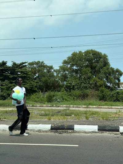 A person stands on the side of a road in Ojomu, Nigeria. The individual, wearing a white t-shirt, black pants, and headphones, holds several balloons. A black jacket hangs over their shoulder. To the side of the road is grassy vegetation. Overhead power lines cross the sky. A curb is painted with black and white stripes.