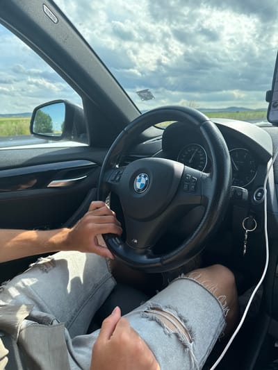 An interior shot of a BMW, taken from the passenger seat looking towards the driver. The driver's hands are on the steering wheel, and the dashboard is visible, including the speedometer and tachometer. The person is wearing ripped denim shorts. The car appears to be driving on a rural road with fields and trees visible outside the windows. The sky is cloudy. The location is not specified as Polsko Padarevo, Bulgaria.