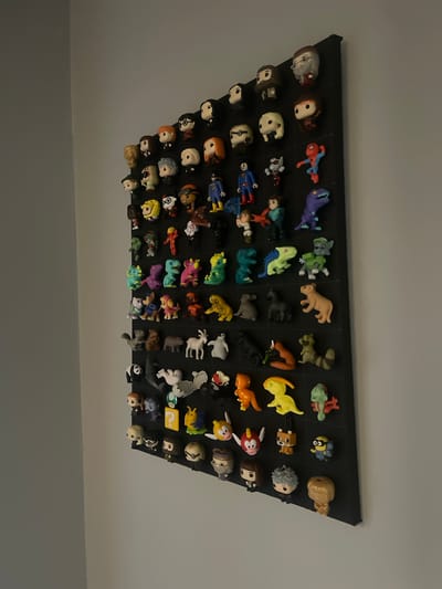A black display board, mounted on a white wall, showcases a large collection of small, colorful figurines. The figurines, arranged in rows and columns, appear to be pop culture characters, including superheroes, cartoon characters, and fantasy creatures. The display board is angled, with the top left corner closer to the camera. The lighting is dim, casting shadows and highlighting the textures of the figurines. The overall impression is one of organized display and collection. This image was taken in Gliwice, Poland.